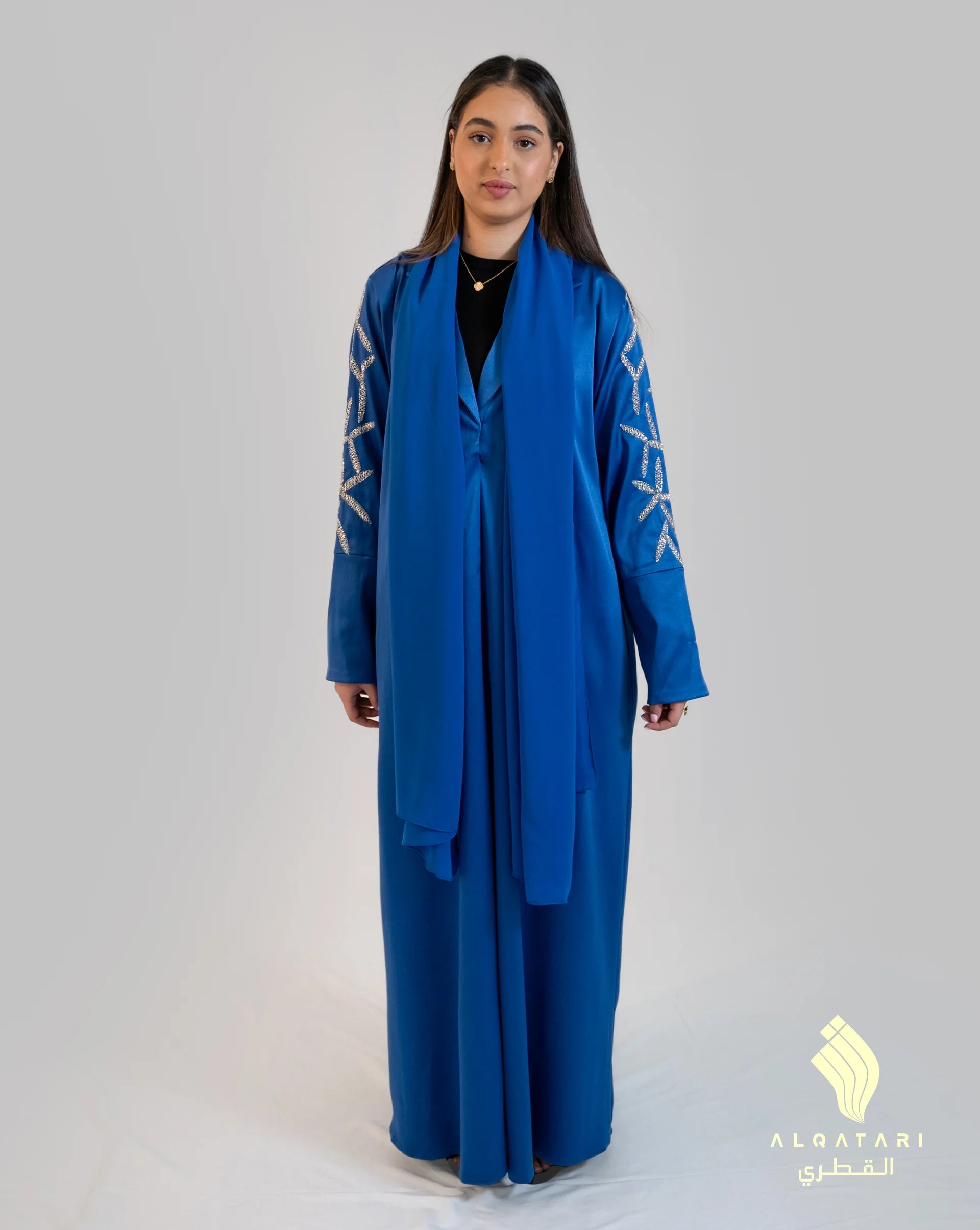 Woman wearing a royal blue abaya with silver embellished sleeve details, standing against a plain light grey background. The abaya is floor-length with a matching scarf and modern modest design by Al Qatari Gallery