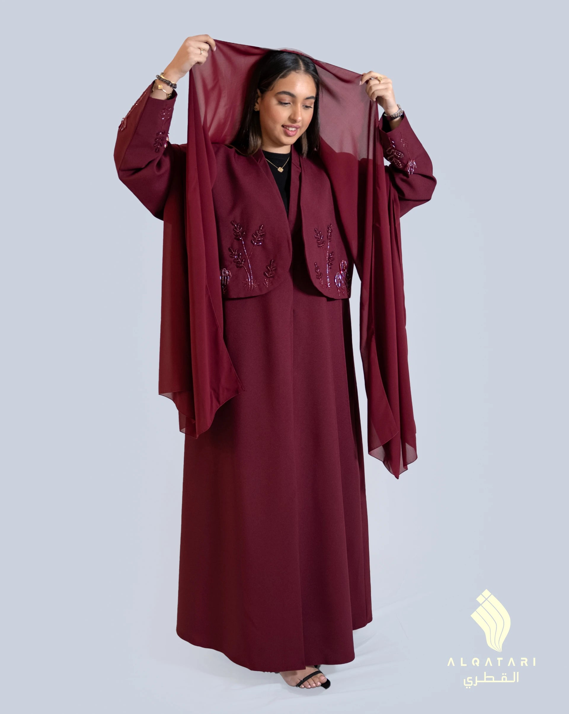 Woman wearing a maroon abaya with a waistcoat-style overlay and delicate floral embellishments, holding up a matching sheer hijab. The modest outfit is elegant and refined, part of the Al Qatari Gallery collection. Available in Brown colour