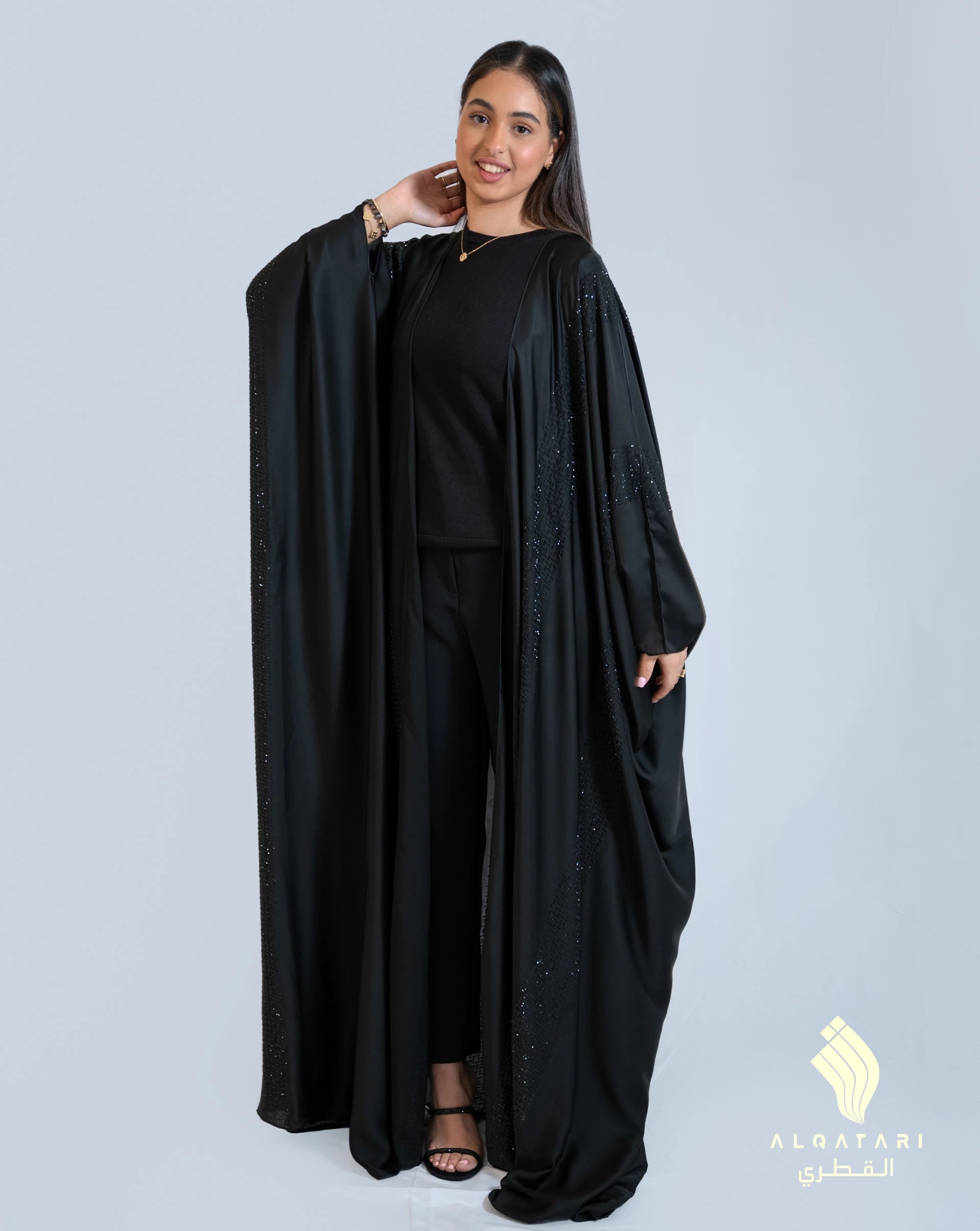 Model wearing a flowing black satin Bahraini abaya from Al Qatari Gallery with intricate black bead detailing along the sleeves and front, showcasing a graceful and elegant look.