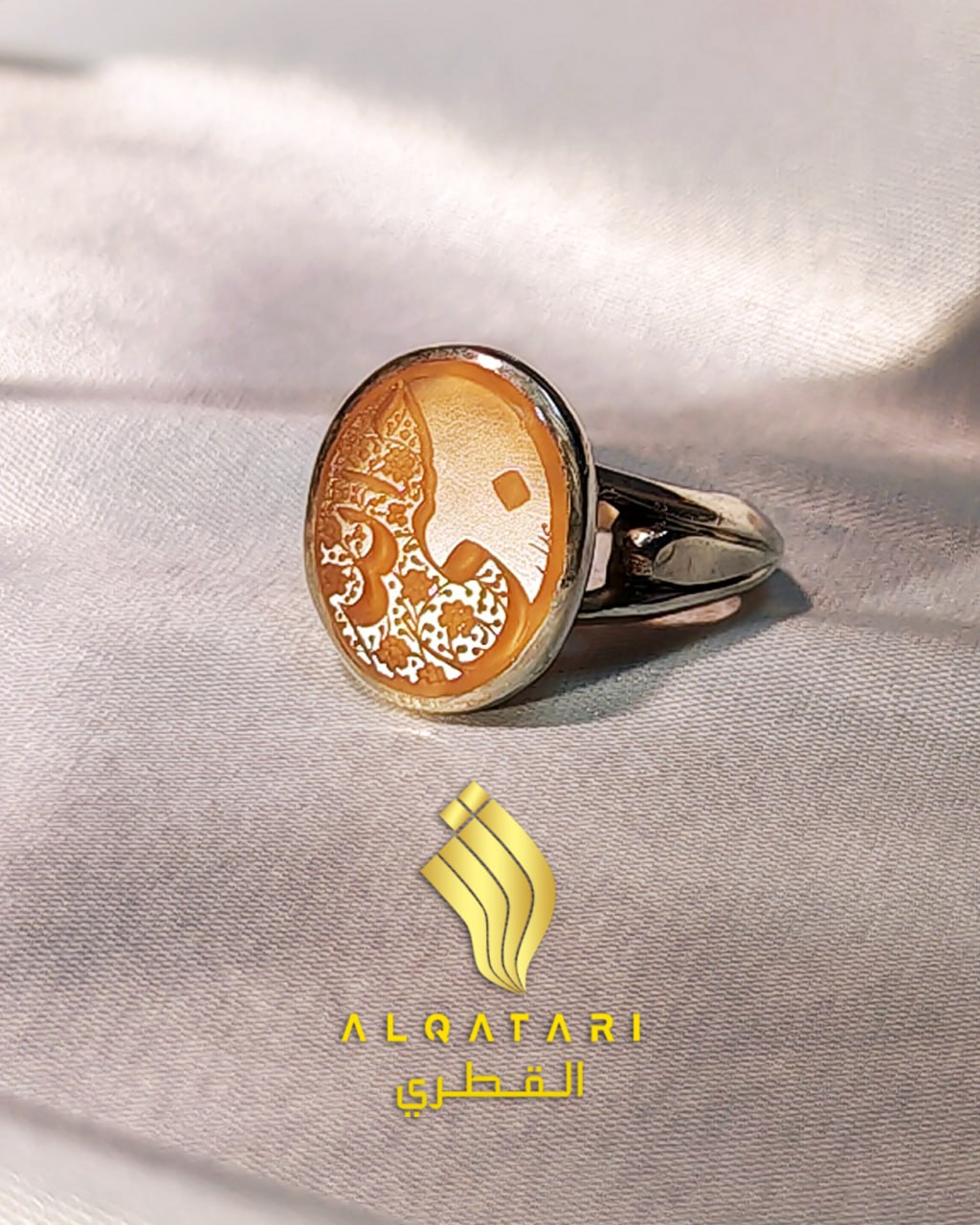 Side view of Al Qatari silver ring with engraved amber agate stone on white satin. Engraving says "Hasbiyallahu" (Arabic: حَسْبِيَ ٱللّٰهُ) In Allah we trustt