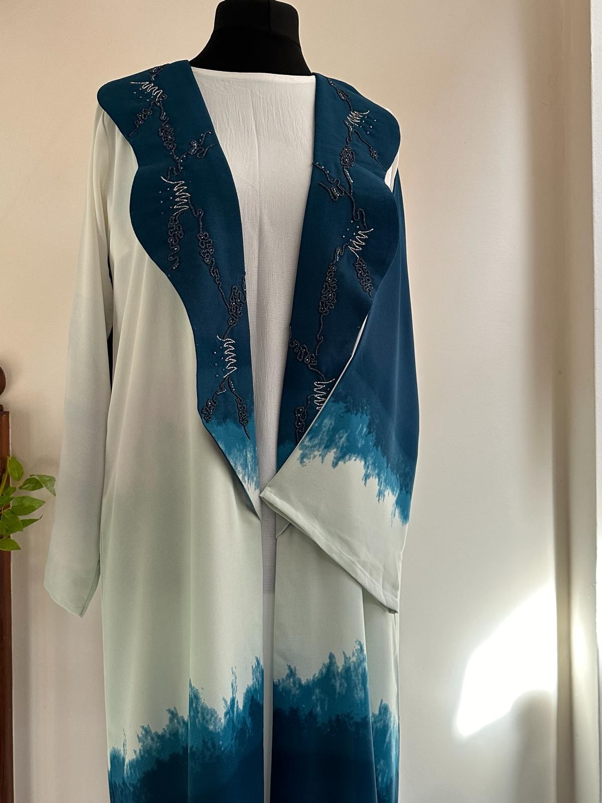Close-up of the blue version of the Waves Ombré Abaya on a mannequin, showing the gradient hem and intricately beaded collar over a white under-dress. Abaya by Al Qatari Gallery Collection